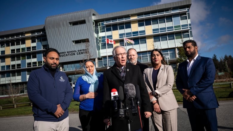 A group of politicians are seen in front of a school making an announcement on a sunny day.