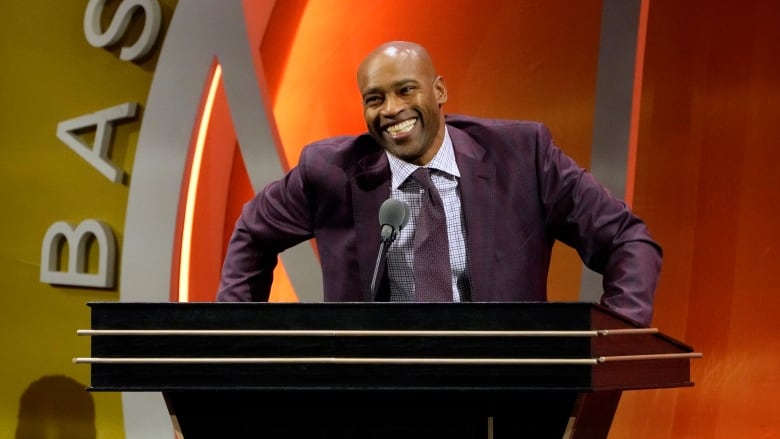 A man wearing a suit and tie smiles while speaking into a microphone at a podium on a stage.