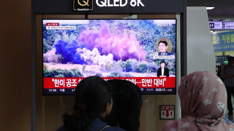 Three people are shown with their backs to camera facing a television in a room that is showing Asian writing and an explosion in a wooded area.