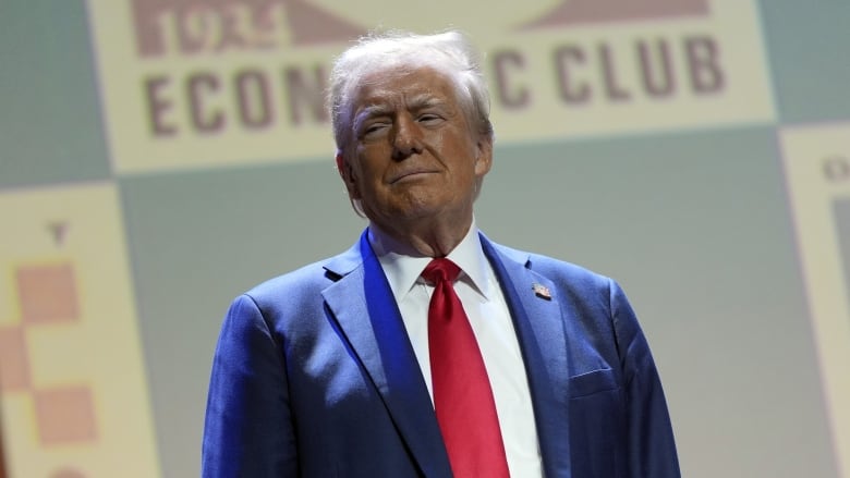 An older cleanshaven man in a suit and tie stands with arms at his side.