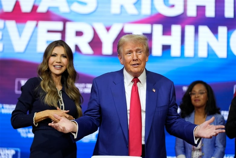 A man in a suit gestures on stage beside a woman