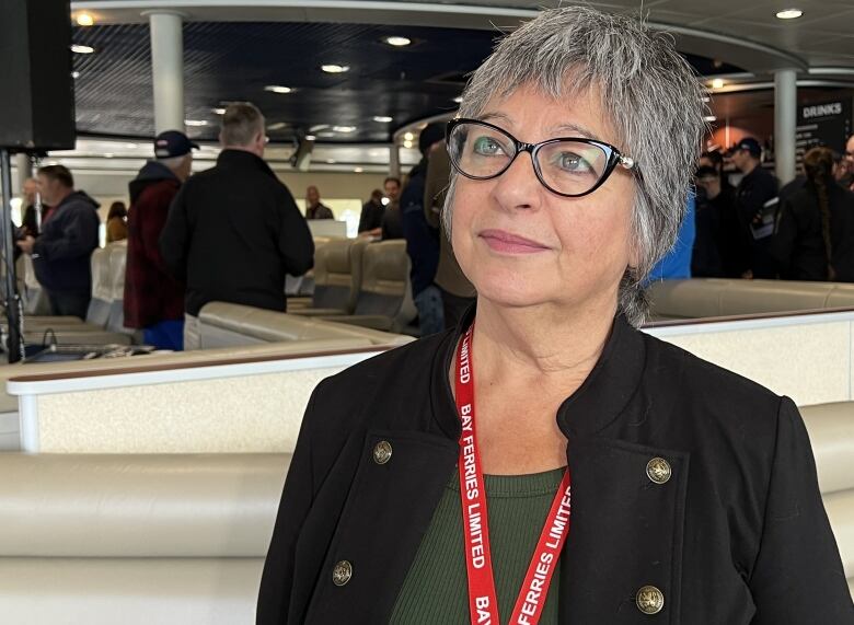 A woman with short grey hair and glasses wearing a blazed and a Bay Ferries lanyard stands on a ferry with people mingling behind her.