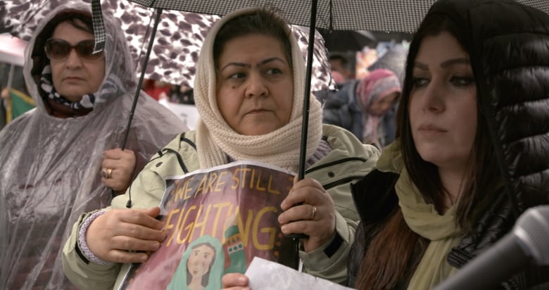 3 woman stand outside, 2 are holding umbrellas. Ayubi, in the middle, holds a sign that says 