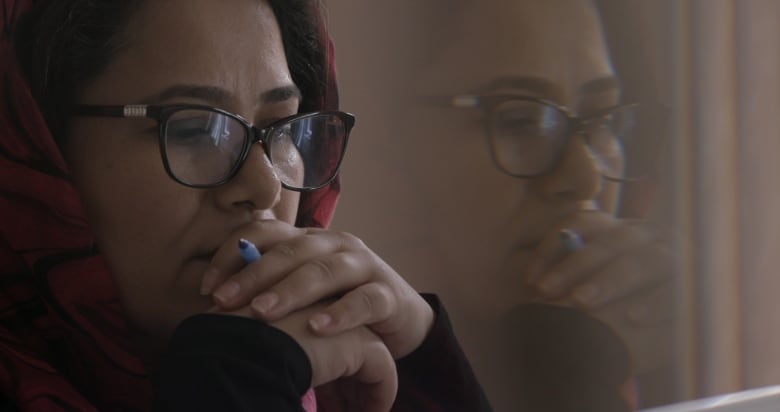 A woman wearing glasses holds her hands in front of her mouth. She is holding a blue pen. Her face is reflected in a pane of glass behind her.