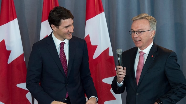 Two men in suits stand together in front of three Canadian flags. One is holding a microphone.