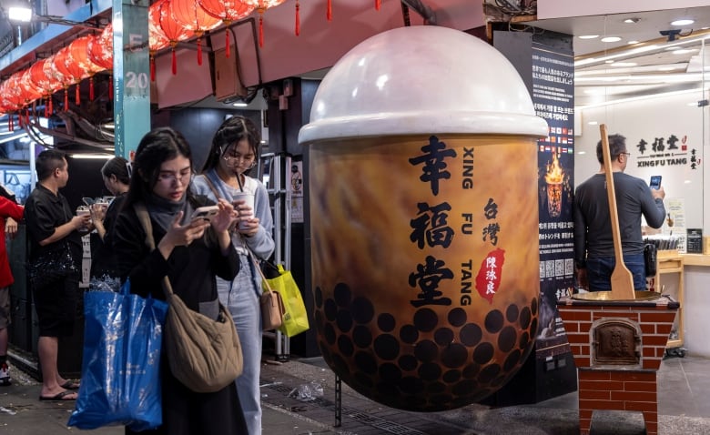 Two women walk past a large bubble tea display in a crowded market. 