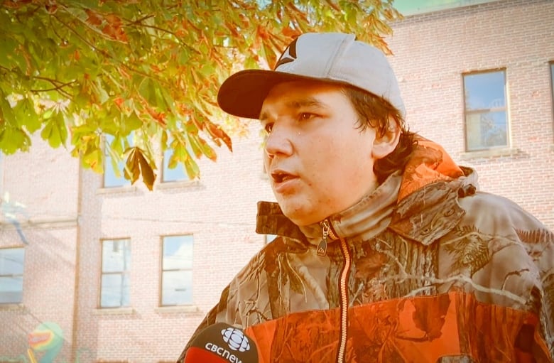  man in an orange patterned camouflage jacket with a grey ballcap speaks with a red brick building in the background.