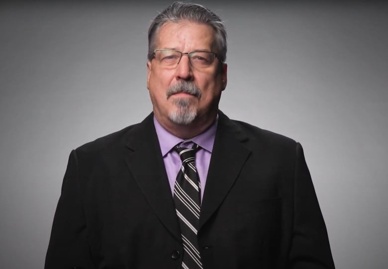 A man stands in front of a grey and white background. They are wearing a black suit over a purple shirt. 