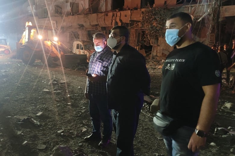 Three men wearing masks over their face stand on the dirt ground near a damaged building in a nighttime photo.