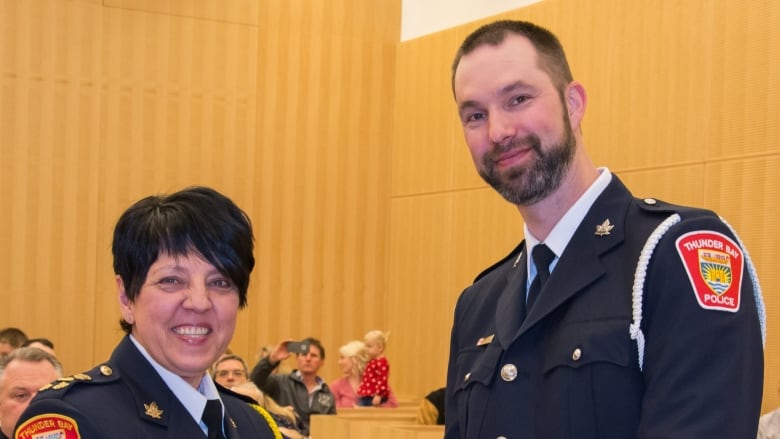 A woman with short hair shakes hands with a tall man with dark hair and a beard. They are in formal police dress and standing in a courthouse.