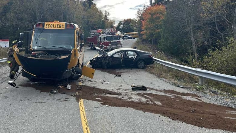 A firefighter standing on a road beside a damaged school bus, with a damaged black car in the other lane, along with spilled fluid and pieces of metal, and a firetruck in the background.