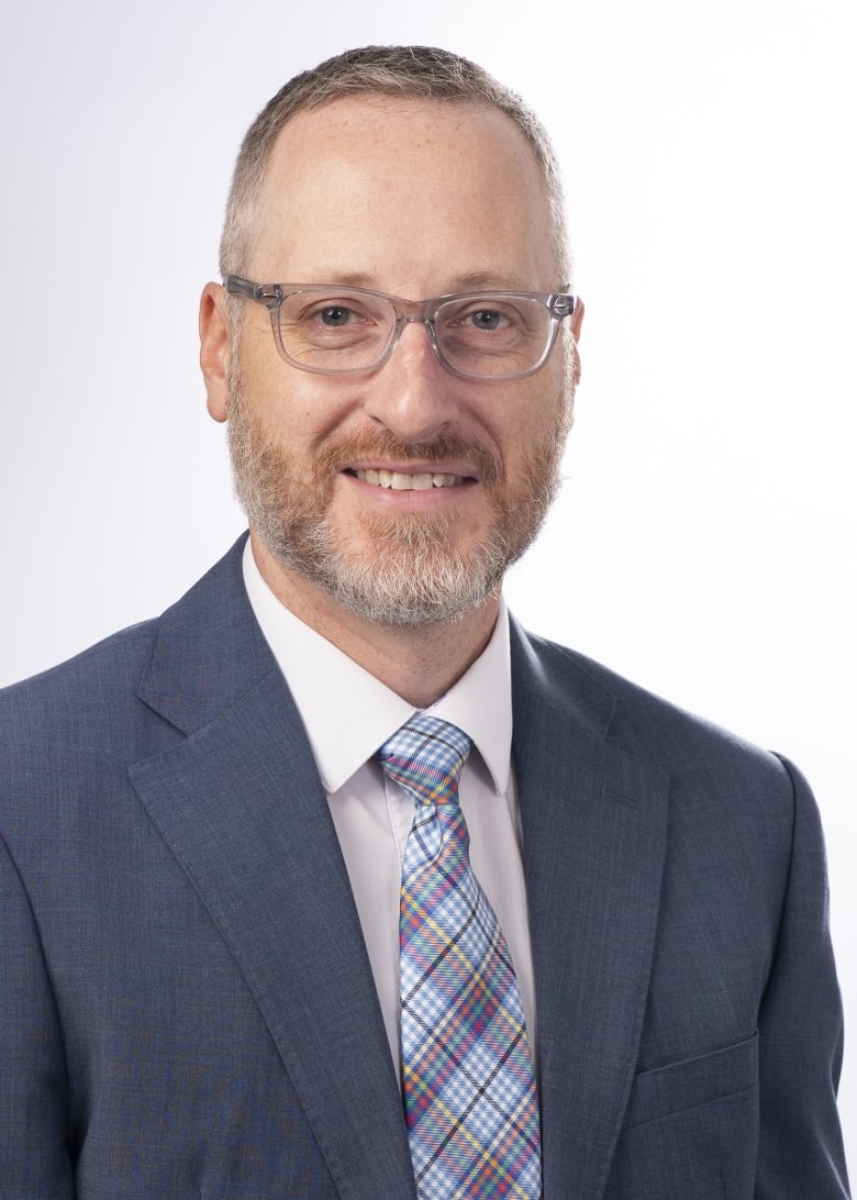 A white man in a dark suit and light shirt and plaid tie has clear-framed glasses and a short beard.