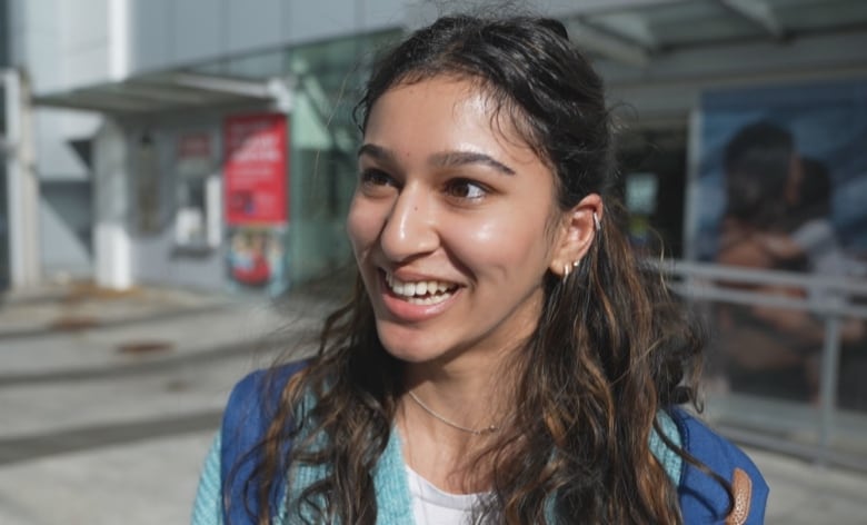 A woman with dark curly hair smiles while standing outside in front of a building.