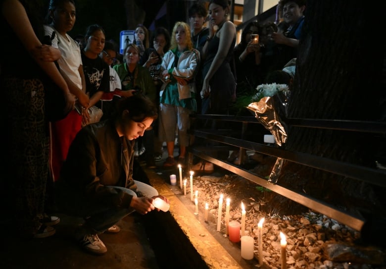 A person crouches to light a candle during a vigil as others crowd around.