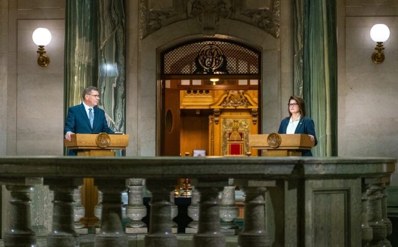 Two people stand at podiums in a legislature building. 