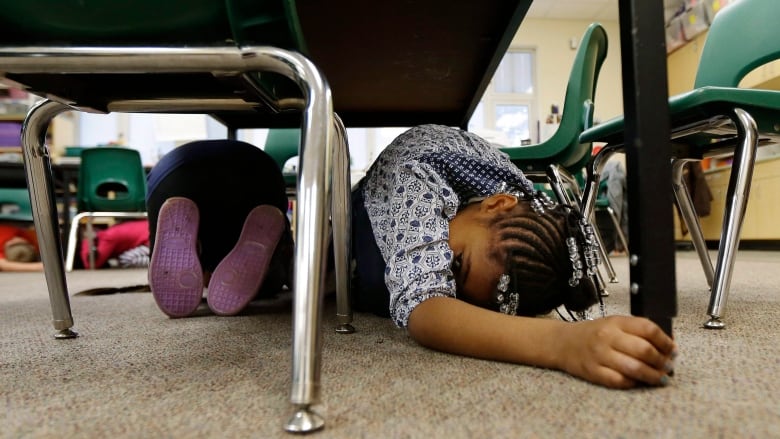 A girl crouches underneath a school table and holds onto the table leg.