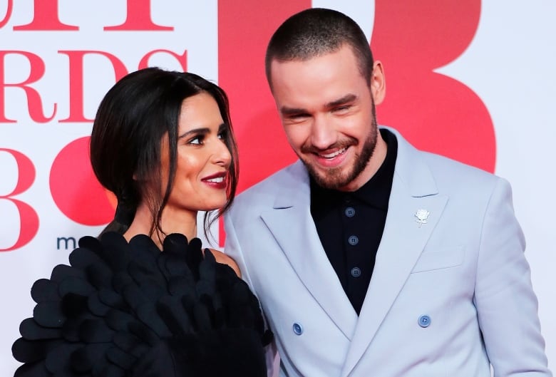 A bearded man in a gray blazer and a woman in a dark dress huddle closely in a red carpet photo.