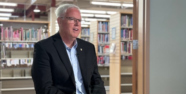 A white man with glasses and white hair wears a black blazer and blue button-up shirt. He sits on a chair in front of an open door, with library shelves behind him