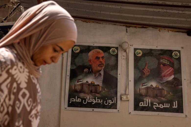 A woman in a headcovering is shown in closeup walking past a wall that has posters of two men.