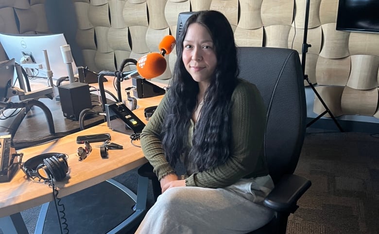 A young woman with long dark hair sits at a table in a radio studio.