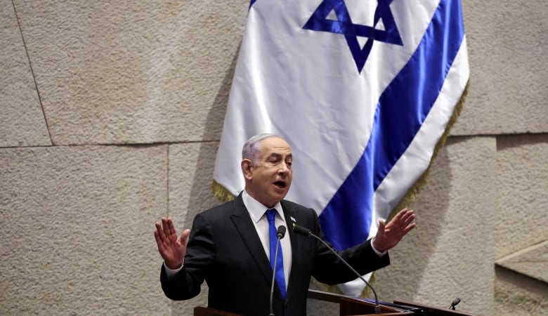 A man gestures while speaking at a lectern. There is an Israeli flag behind him. 