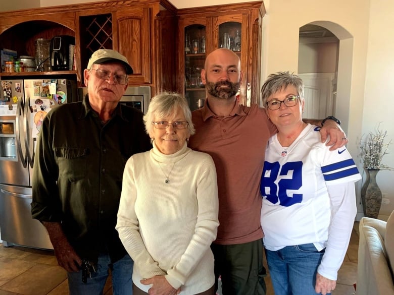 Two men and two women stand in a kitchen with their arms around each other's shoulders.