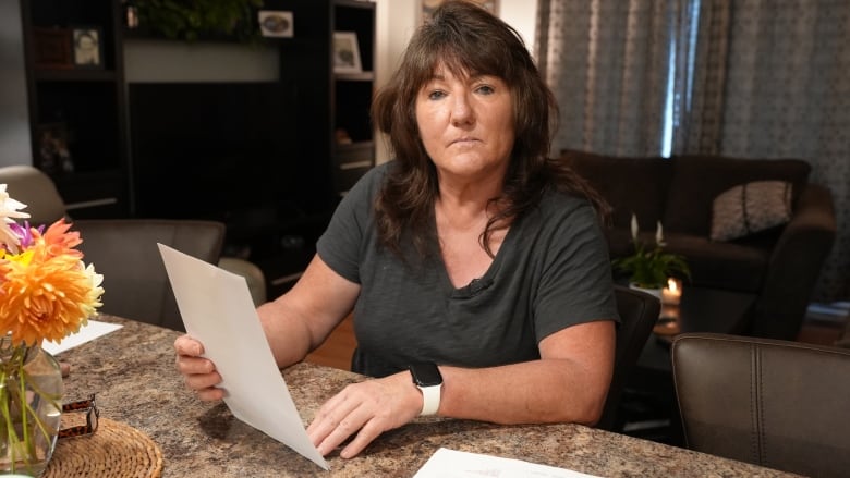 A woman sits at a table in a living room, holding a piece of paper. She doesn't look happy. 