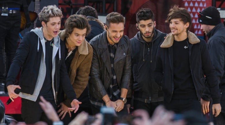 Five men laugh together in a line, most looking down towards a crowd. There are blurry hands of the crowd visible at the bottom of the image. The men are wearing jackets or hoodies. 