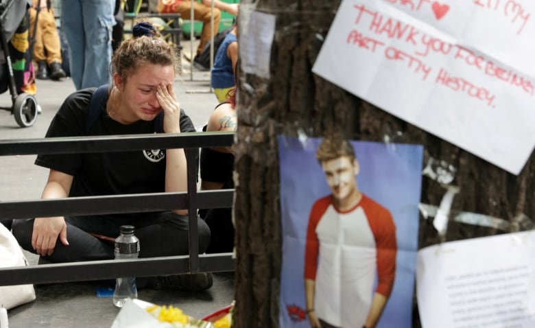 A woman sits cross-legged on the pavement, crying. In the blurry foreground is a photo of Liam Payne on a wall or tree trunk (it's unclear which) with other signs mourning him. 