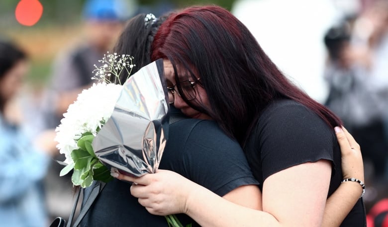 Two women hug each other, one holding a bundle of white flowers. 