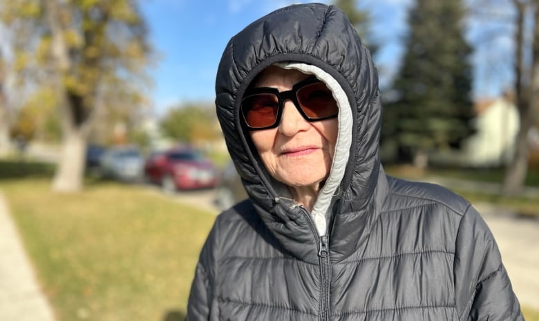 An elderly woman is standing outside, wearing a black jacket with the hood pulled up, and sunglasses covering her eyes.