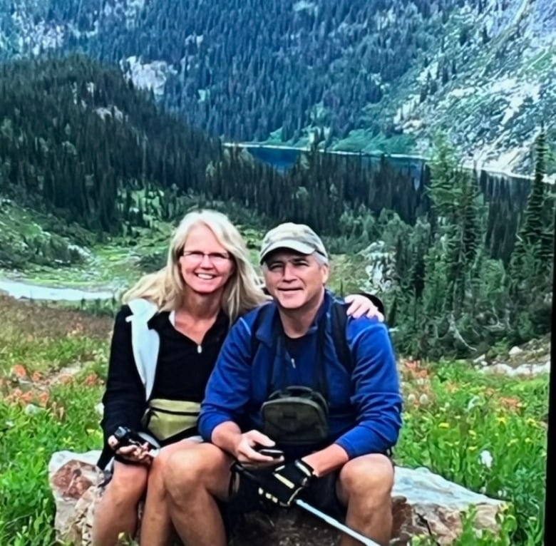 Woman and man seated on a rock in front of a lake and mountain scene. 