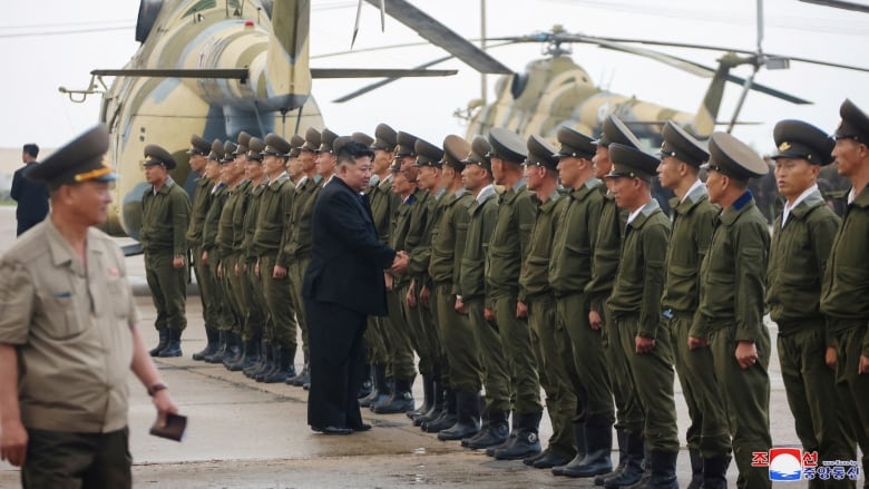 A line of Asian men in military uniforms is shown, with a man in a suit shaking the hands of one soldier.