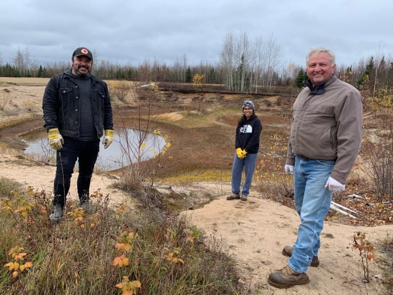 Three people, two men and one woman, are standing at a sandy ground, and near them is a small pond.