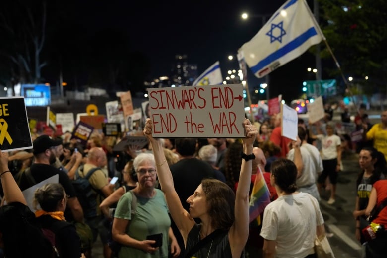 Dozens of people are shown in a nighttime photo, some holding signs and an Israeli flag in the background. A woman in the holds a handmade sign that says 'Sinwar's end, end the war.'