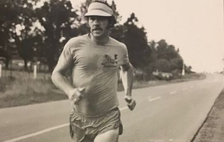 Moustached man in sweaty t-shirt, visor, shorts runs along paved road.