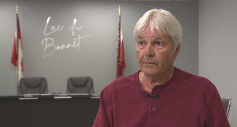 A man with white hair and a red shirt sits in front of some flags and a Lac du Bonnet logo on the wall.