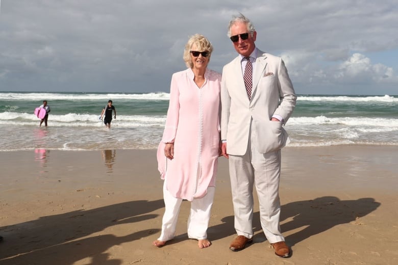 Two people wearing sunglasses stand on a beach with two surfers walking in the water behind them.