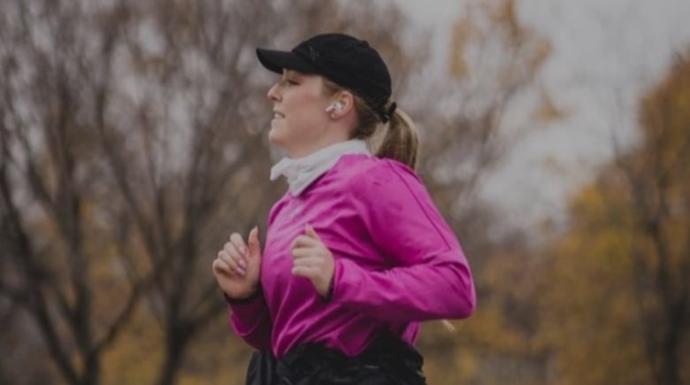 A woman in a pink jogging suit and black ball cap, with an airpod in her ear, jogs on a grey street on a cold fall day