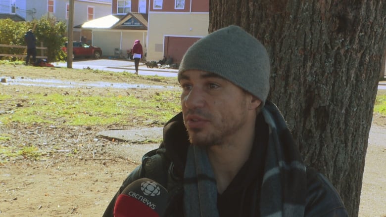 A man in a grey beanie sits in front of a tree.