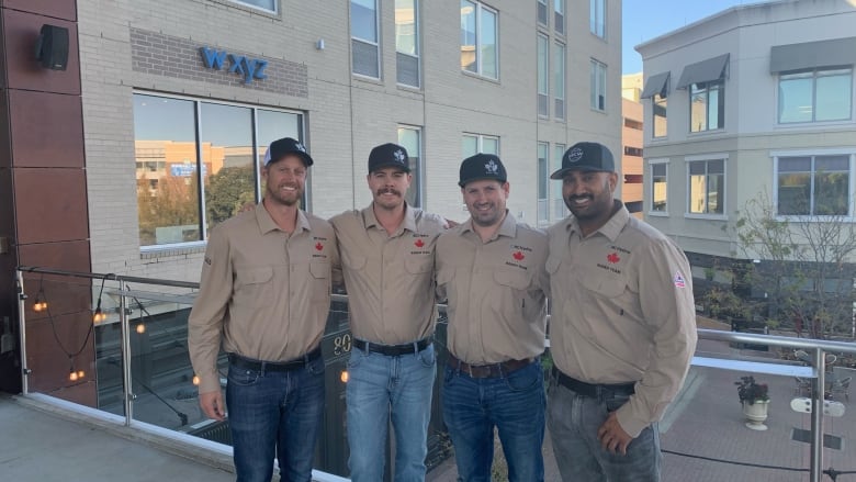 Four men in matching jeans, beige work shirts ad blue caps stand smiling with their arms around each other's shoulders.