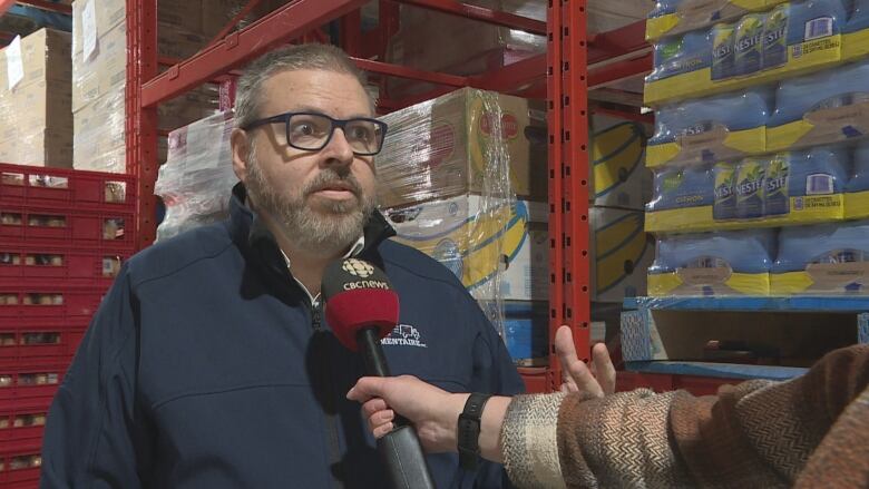 A man in a blue jacket stands in a food bank with walls full of boxes of food.