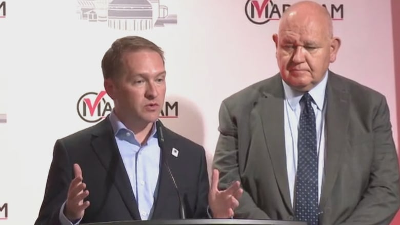 Two men in suits, one bald and the other with blonde hair, stand at a podium. The background behind them says 