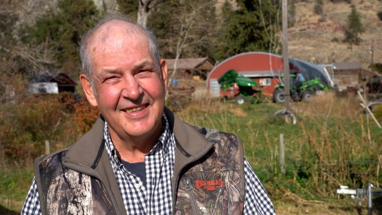 A profile photo of a smiling man with a checkered shirt and camouflaged vest standing in a field with his farm equipment and building in the backgroud. 