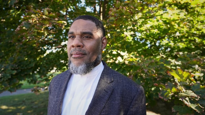 A man in a white shirt and grey suit jacket is pictured in front of a green leafy tree.