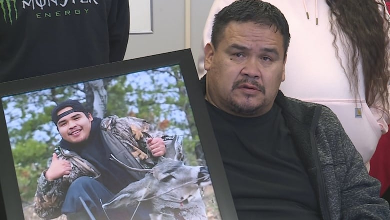 A man in black holds up a photograph of a young man. 