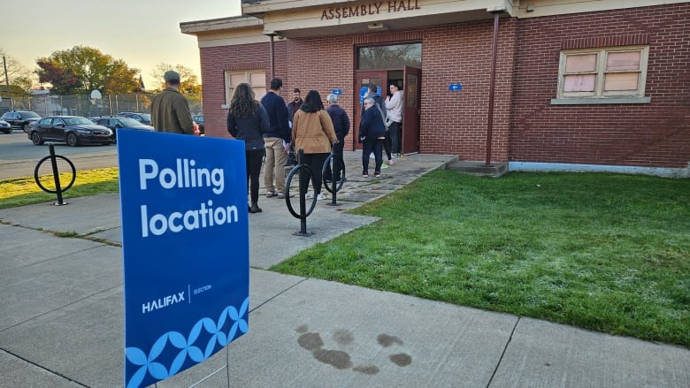 A group of people can be seen waiting in line outside Assembly Hall. In the foreground, a blue sign advertises this hall as a polling location.