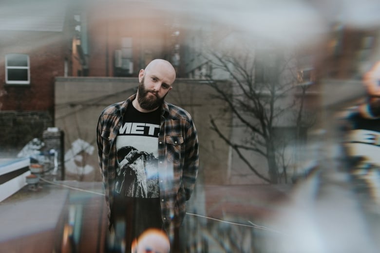 A man stands on a rooftop. He is bald and has facial hair, and is wearing a plaid shirt over a tee shirt that says 