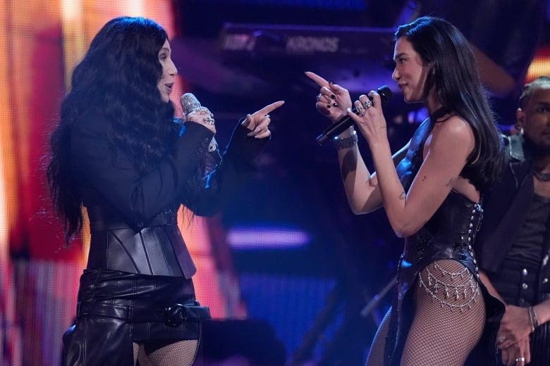 Two pop stars sing into mics while wearing black leather and fishnet stockings.