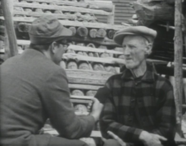 A black and white photo of two men sitting in front of a wood pile. 